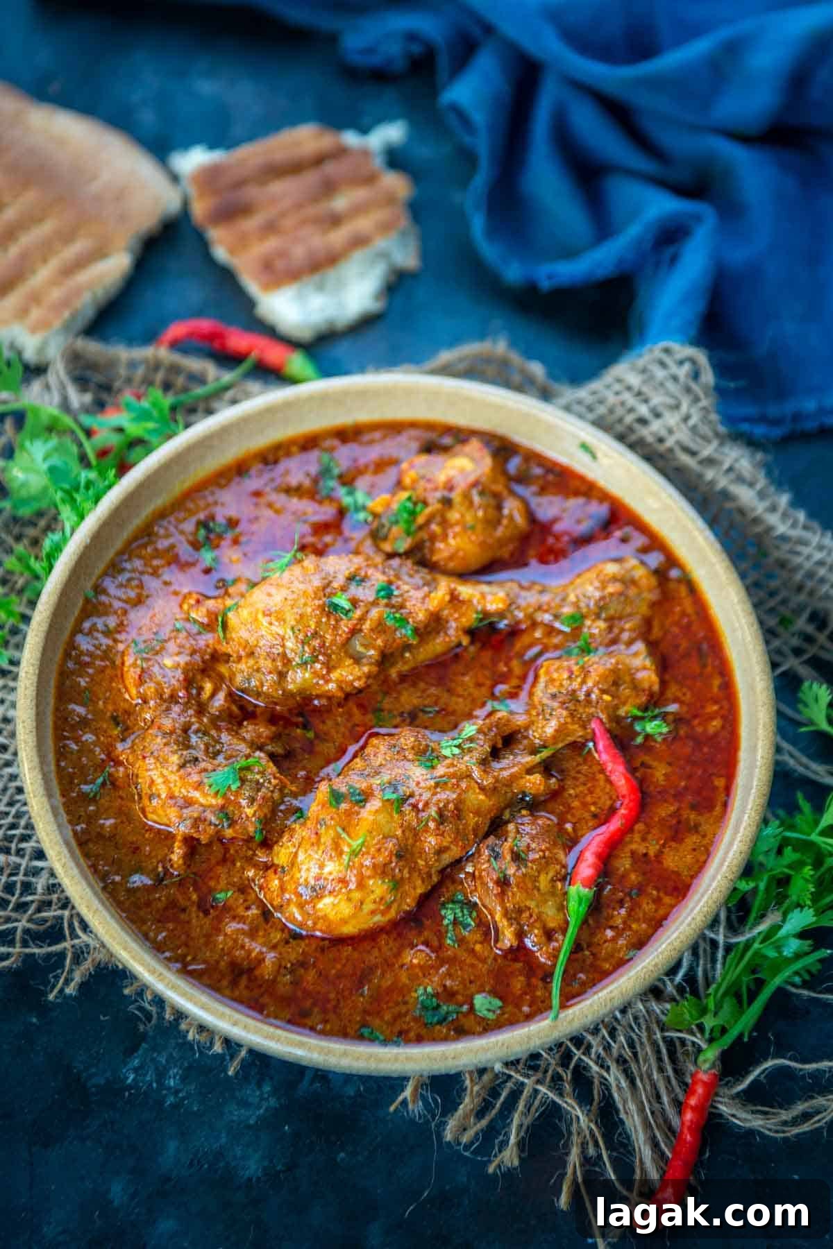 Chicken Angara Recipe: Spicy Grilled Chicken with Smoky Sauce 2 A bowl of spicy Chicken Angara curry garnished with fresh herbs and red chilies, surrounded by pieces of flatbread and a dark blue cloth on a rustic surface.