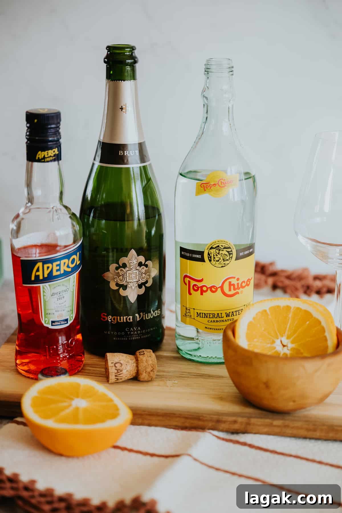 Classic Aperol Spritz Recipe: How to Make the Perfect Cocktail 3 bottles of Aperol, Segura Viudas Cava, and Topo Chico on a wooden surface with a bowl of orange rounds and a wine glass.