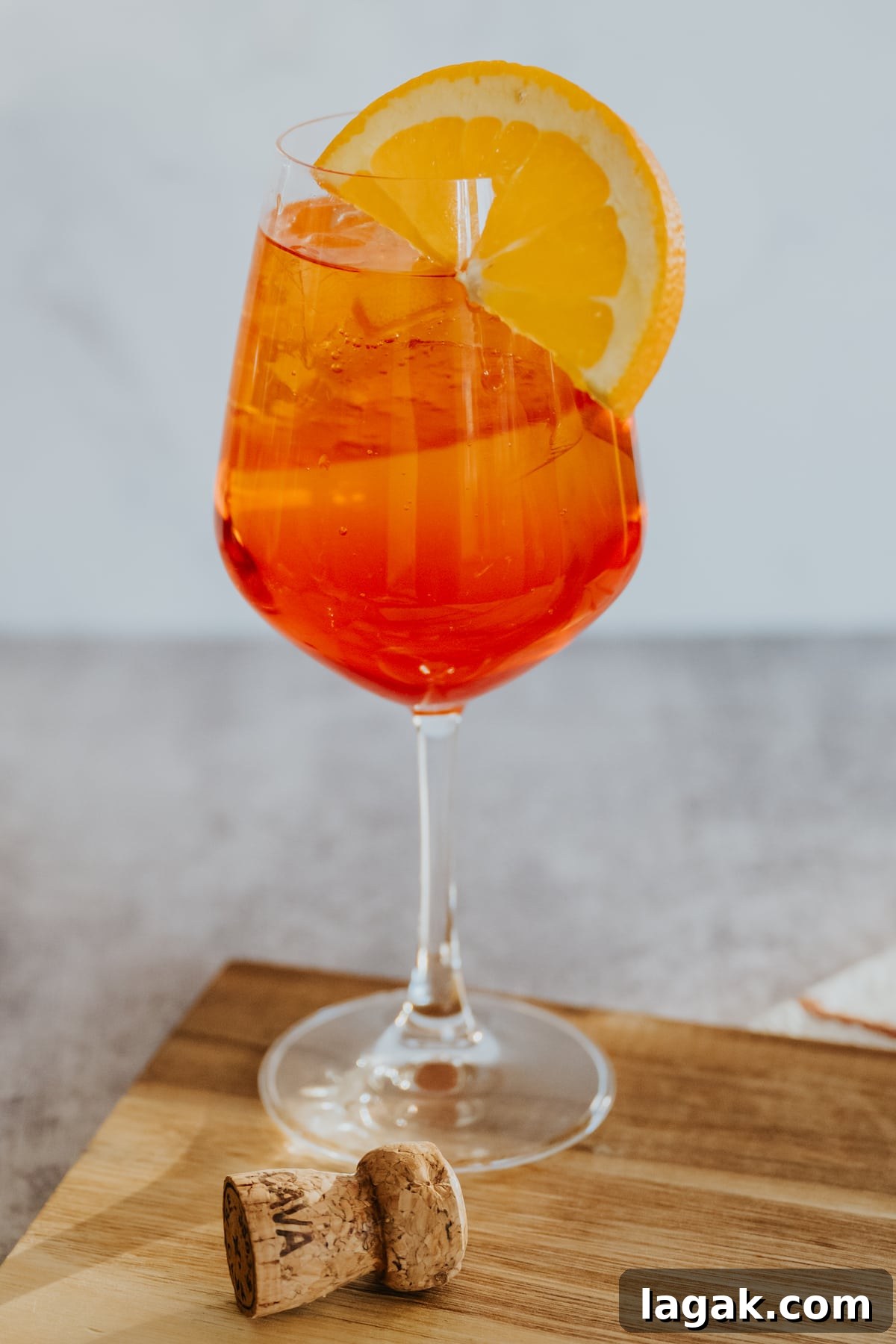 Classic Aperol Spritz Recipe: How to Make the Perfect Cocktail 7 stemmed white wine glass filled with an Aperol spritz over ice garnished with an orange slice on a wooden cutting board with a cava cork bottle in the foreground.