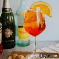 Classic Aperol Spritz Recipe: How to Make the Perfect Cocktail 10 hero shot of a stemmed wine glass filled with an Aperol spritz on ice garnished with an orange round in front of a cava and topo chico bottle.