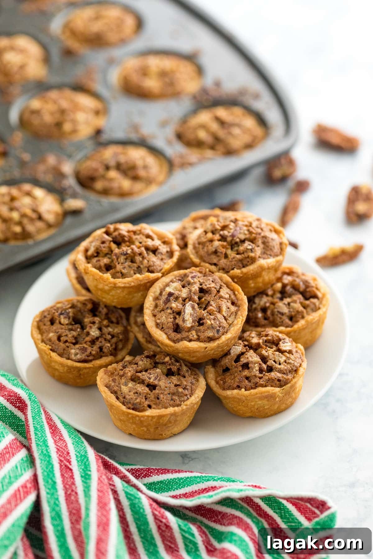 Grandma Rose’s Pecan Tassies Recipe for Perfect Bite-Sized Pies 2 A plate of mini pecan tarts with a mini muffin tin of them in the background, a red and green striped towel and some broken pieces of it.