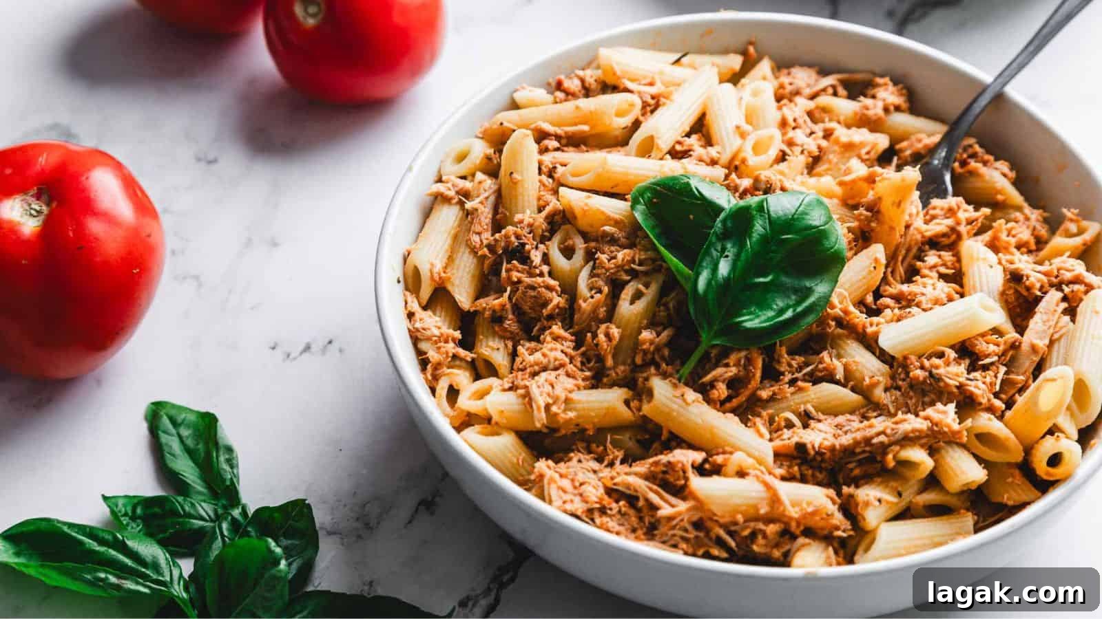 21 Slow Cooker Dinners for Stress-Free Weeknight Meals 19 A bowl of penne pasta topped with shredded meat and garnished with fresh basil leaves. A fork is placed in the bowl. Red tomatoes and additional fresh basil leaves are on the marble surface nearby.
