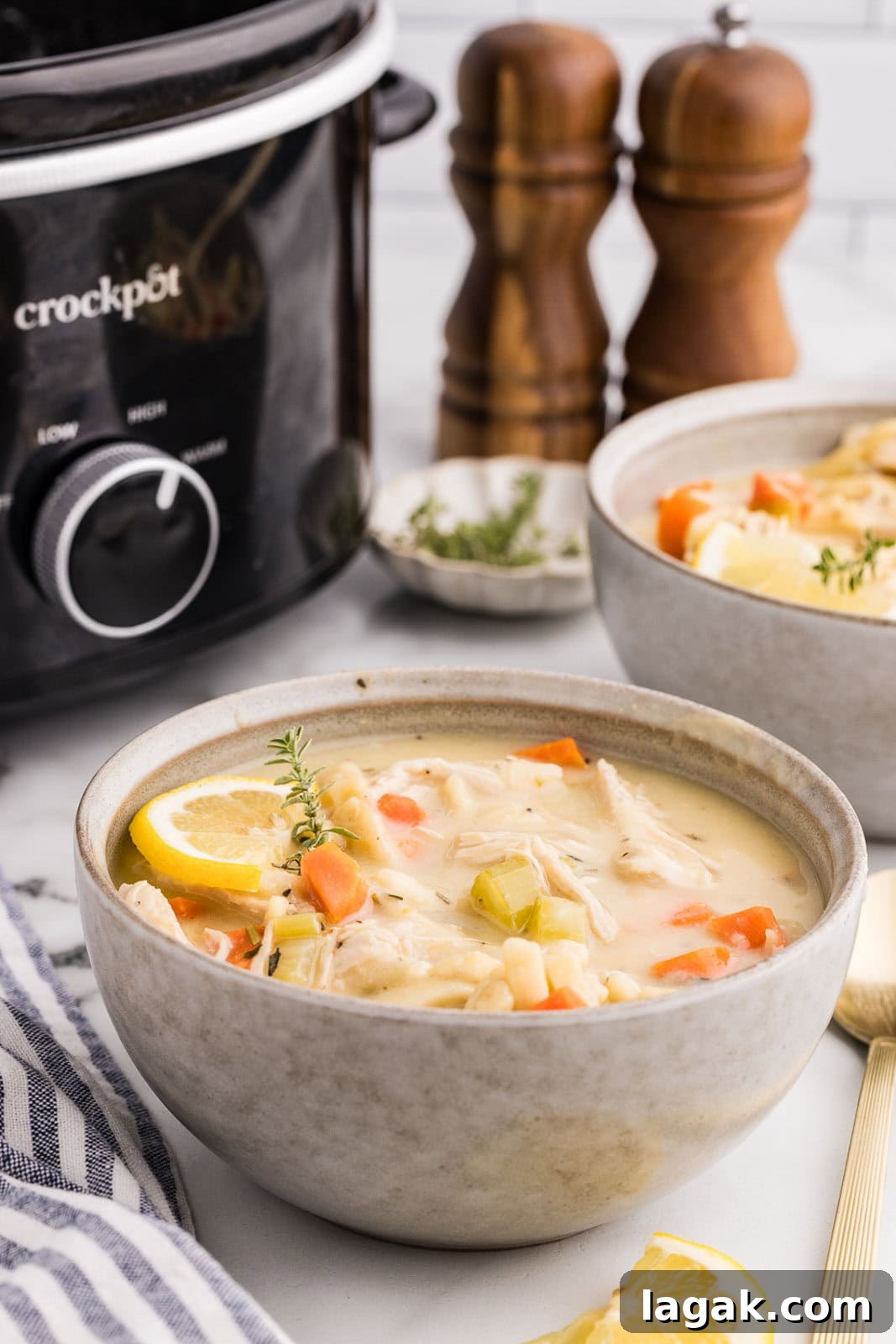 side view of bowl of chicken noodle soup surrounded by wooden salt and pepper shakers, another bowl of soup, gold spoons, and black slow cooker