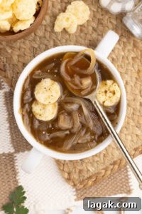 overhead view of white soup cup of onion soup and Parmesan crisps with silver spoon on a tan place mat with bowl of Parmesan crisps and salt and pepper shakers in the background
