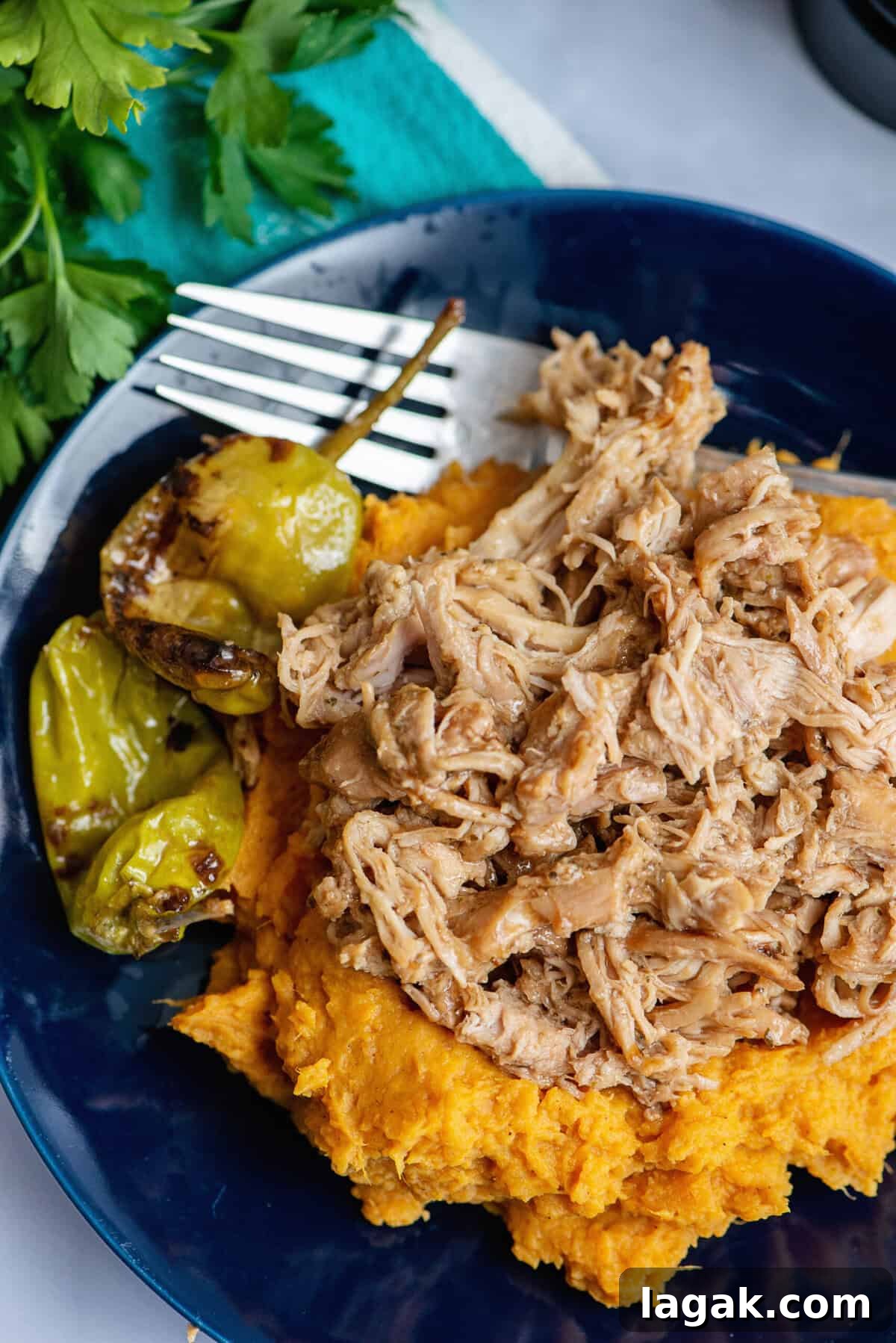 Mississippi Chicken Served Over Creamy Mashed Sweet Potatoes 2 Mississippi chicken served over a generous bed of mashed sweet potatoes, garnished with pepperoncini peppers, all presented on a dark blue plate with a fork and fresh parsley nearby.