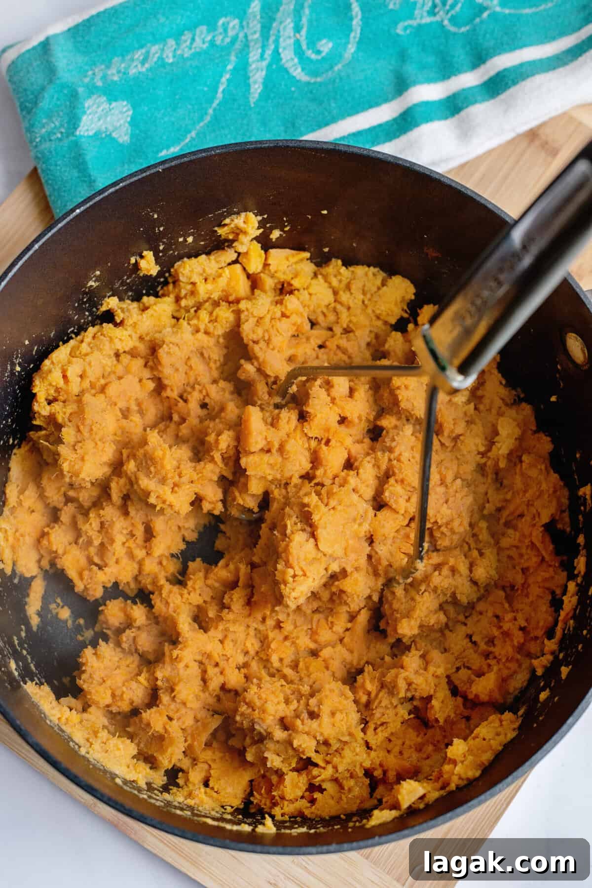 Mississippi Chicken Served Over Creamy Mashed Sweet Potatoes 13 Pot of mashed sweet potatoes being blended with a handheld masher, showing a fluffy, orange texture, with a teal kitchen towel and wooden cutting board in the background.