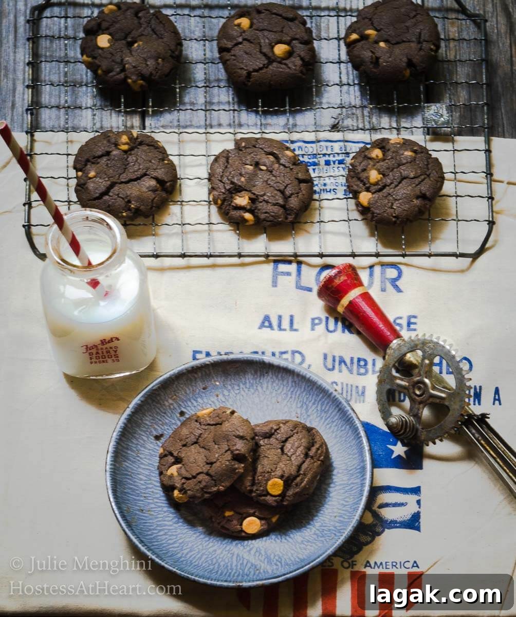 No-Chill Dark Chocolate Peanut Butter Cookie Recipe 4 Top-down photo of Chocolate cookies dotted with peanut butter chips sit on a cooling rack over a flour sack. A blue plate holding two cookies is in the front next to a jar of milk and an antique hand mixer sit to the side.