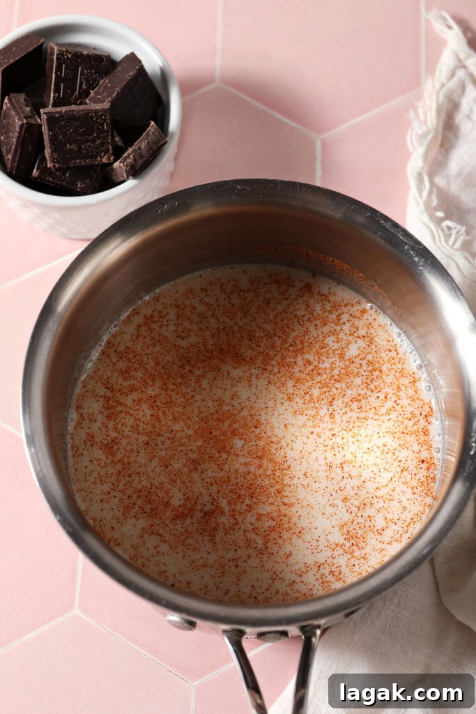 Overhead of a saucepan holding milk and cayenne pepper on a pink surface near a bowl of chocolate