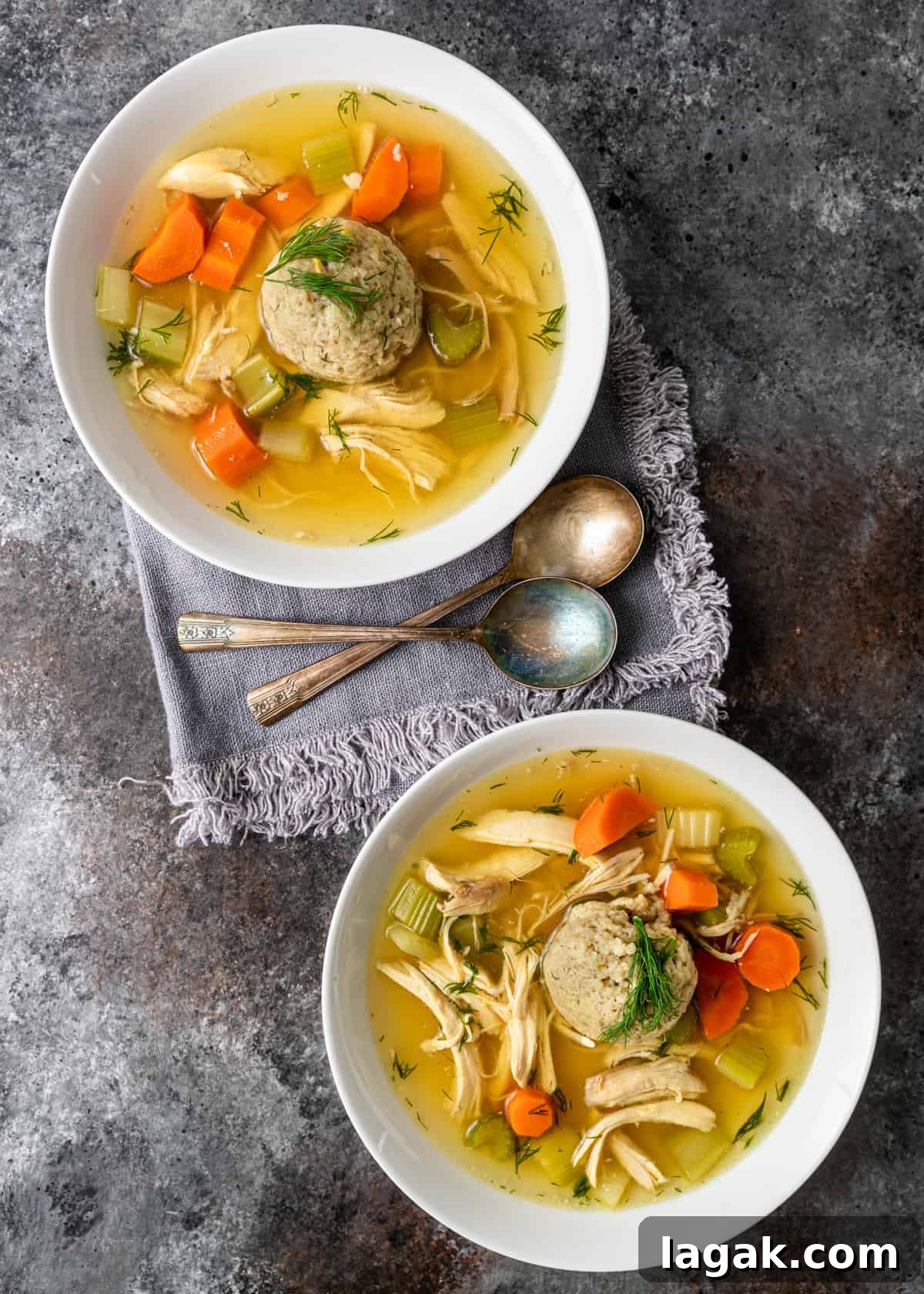 overhead: matzo ball soup in two white bowls with two spoons on gray linen