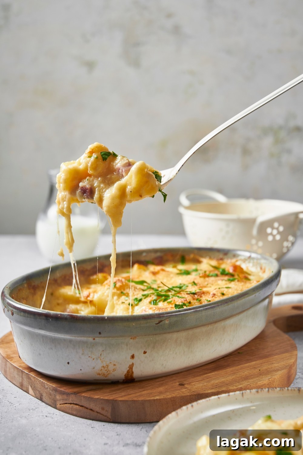 Casserole dish on a wooden board with a scoop of ham and potato casserole being scooped out of the dish by a large serving spoon.