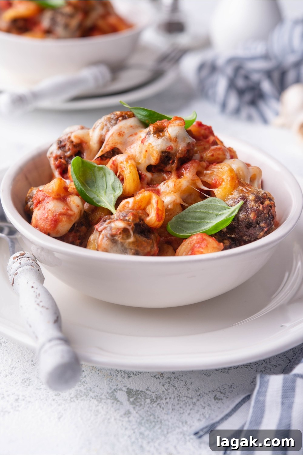 A white bowl of meatball casserole on a white plate with a fork on the plate. In the background there is another serving of casserole.