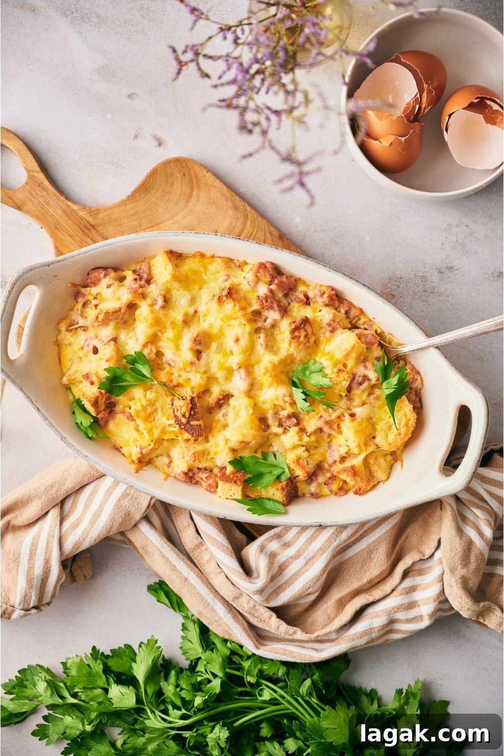 Christmas breakfast casserole in a white casserole dish with a serving spoon in the dish. The dish is on a wood board with a kitchen towel next to the dish.