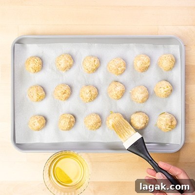 hand holding pastry brush with black handle, brushing yellow olive oil on raw chicken meatballs